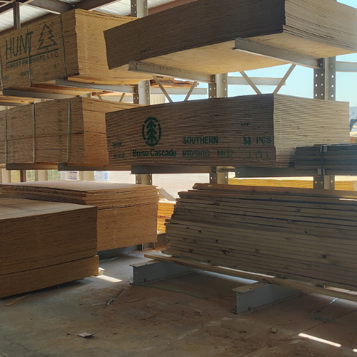 Stacks of plywood and lumber sit on metal racks in a sunlit building-supply warehouse, with branded wood panels and an industrial feel.