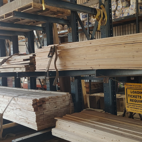 Stacks of wooden lumber fill industrial warehouse shelving, with hanging straps and a yellow “loading tickets required” sign.