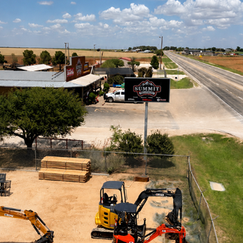 Rural equipment yard with excavators, lumber stacks, and a Summit sign beside a highway, surrounded by fields under a bright, cloudy sky.