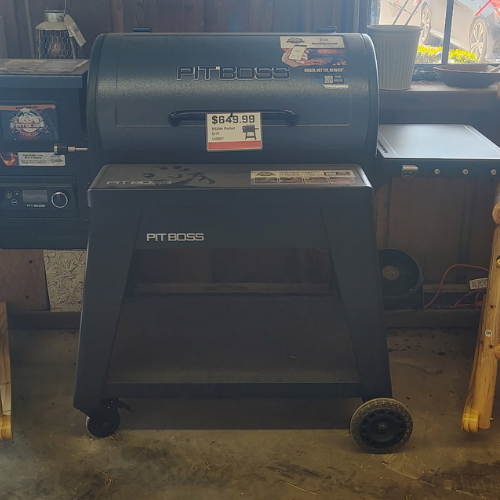 Pit Boss black pellet grill on wheels displayed indoors, with a $649.99 price tag, control panel, chimney, and rustic workshop setting.