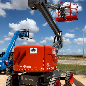 Bright outdoor equipment yard with a red Skyjack telescopic boom lift raised high, blue lift behind, chain-link fence, and cloud-filled sky.