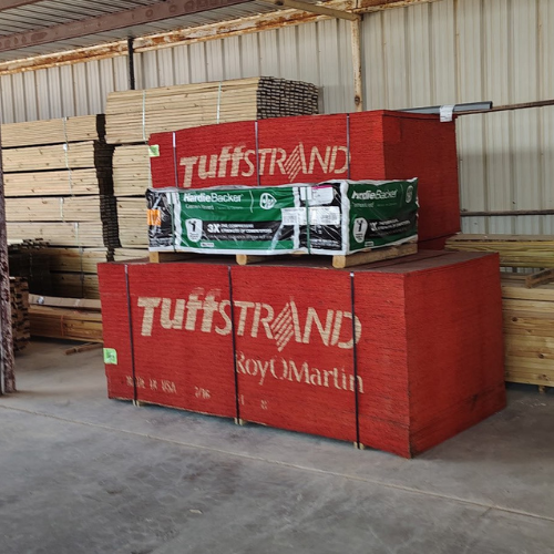 Stacked lumber bundles in a warehouse, including red Tufstrand and green HardieBacker-wrapped materials, conveying an industrial, organized setting.