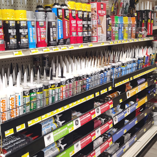 Hardware store shelf stocked with colorful caulk, sealants, construction adhesives, and filler tubes, creating a busy, well-organized retail display.