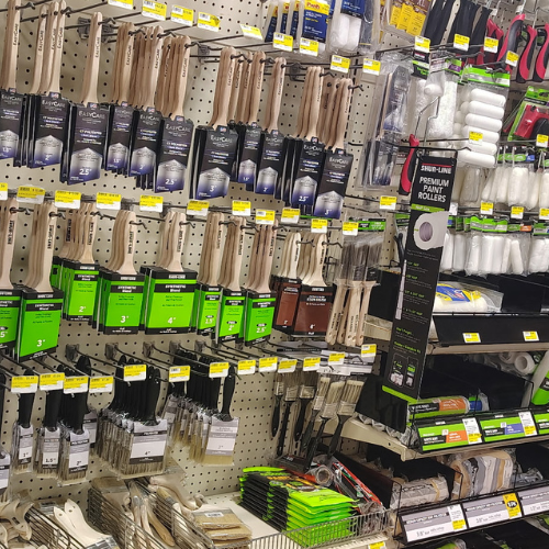 Hardware store aisle displaying rows of paintbrushes, rollers, and painting tools on pegboards and shelves, with colorful labels and packaging.